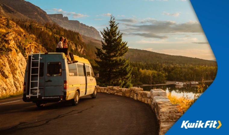 Two people sit on the roof of a camper van, overlooking a beautiful lake at sunset.