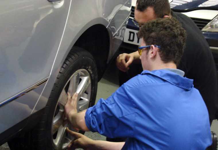 Kwik Fit apprentice is instructed by a trainer on how to change a tyre on a car.