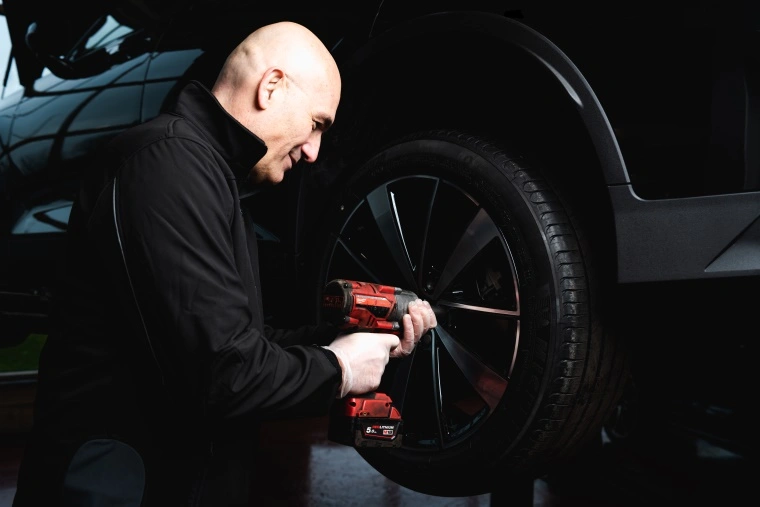 Man in a black jacket with a red power tool fitting a new tyre to a black car