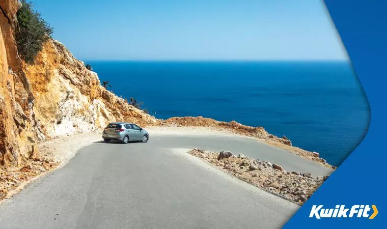 A steep mountain road in Greece contrasts with the shockingly blue sea and sky in the distance.