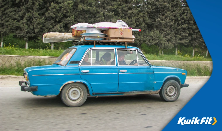 A family drives in a charming, boxy old blue car with a loaded roof deck.