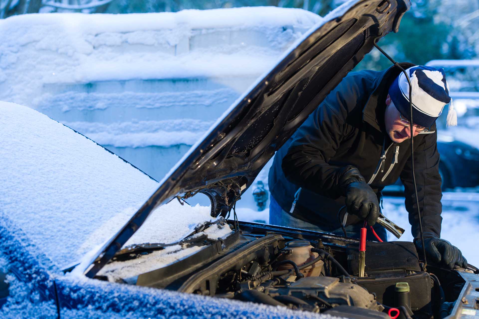 Man wearing a hat and gloves attaching jump leads to a car battery in the snow.