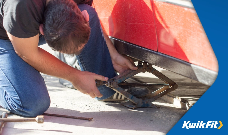 A man uses a scissor jack to raise his red car off the ground enough to replace a flat tyre.