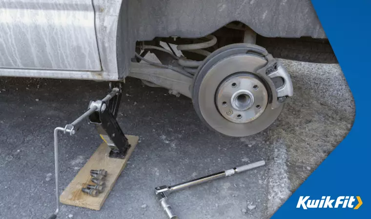A jack lifts up a car while the wheel has been removed with a lug wrench to reveal the calliper.