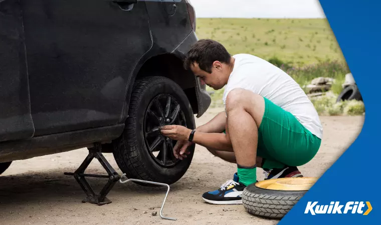 A driver in a white shirt and green shorts is replacing a punctured tyre with their spare wheel.