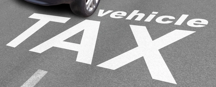 The words 'Vehicle Tax' are written on the road as markings while a white car drives over them in the top left corner.