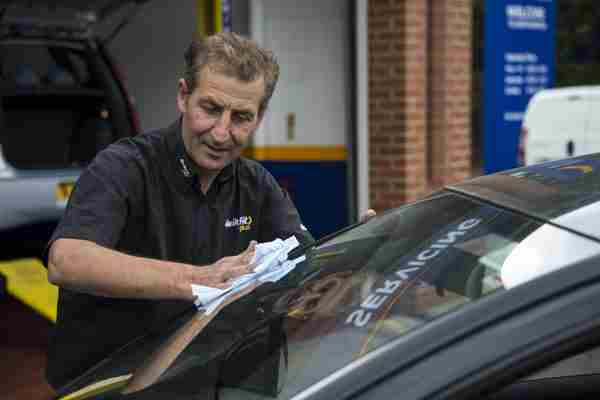 A Man in a Kwik Fit top wiping a windscreen of a car during an MOT test